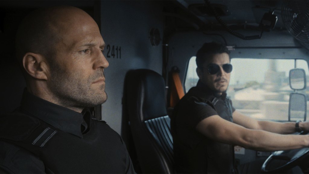 This is an image of two middle aged men in the front seats of some kind of vehicle. The man driving is wearing sunglasses and is looking to the man in the passenger seat who is looking forward with a blank expression on his face. 